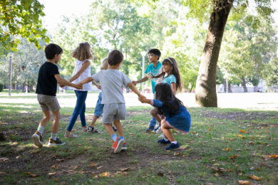 Kinder spielen im Kreis auf einer grünen Wiese im Park. Bäume im Hintergrund.